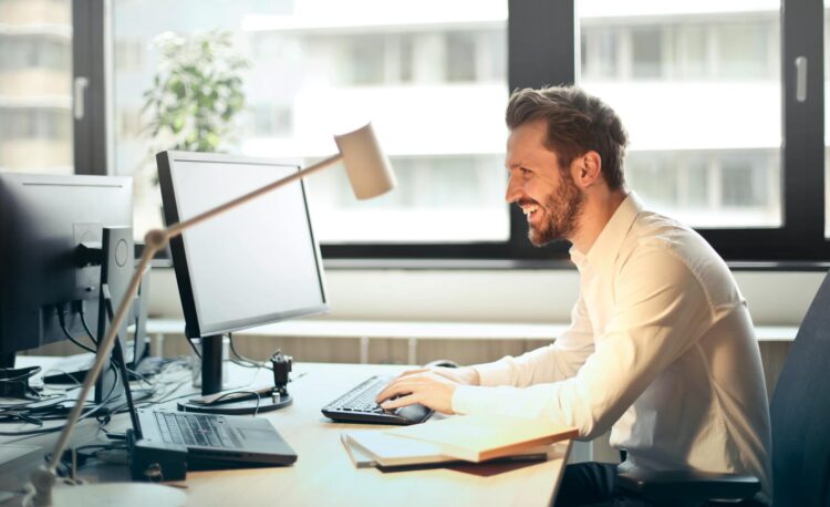 man in white dress shirt sitting on black rolling chair while facing black computer set and smiling