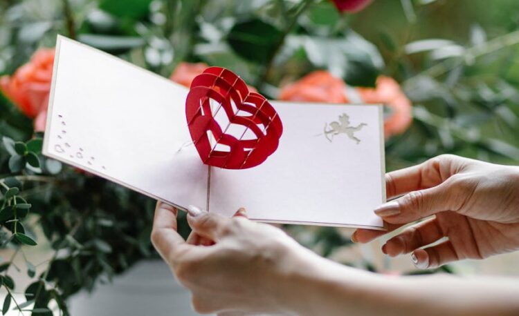 crop woman with valentine card near blooming flowers